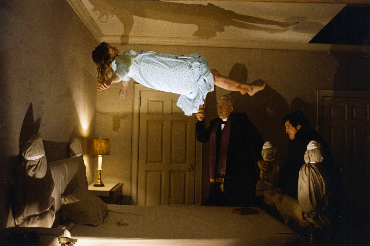 <엑소시스트>Regan McNeil (Linda Blair) levitates above a bed in front of Damien Karas (Jason Miller) and Father Lancaster Merrin (Max von Sydow).” class=”wp-image-905916″/><button class=