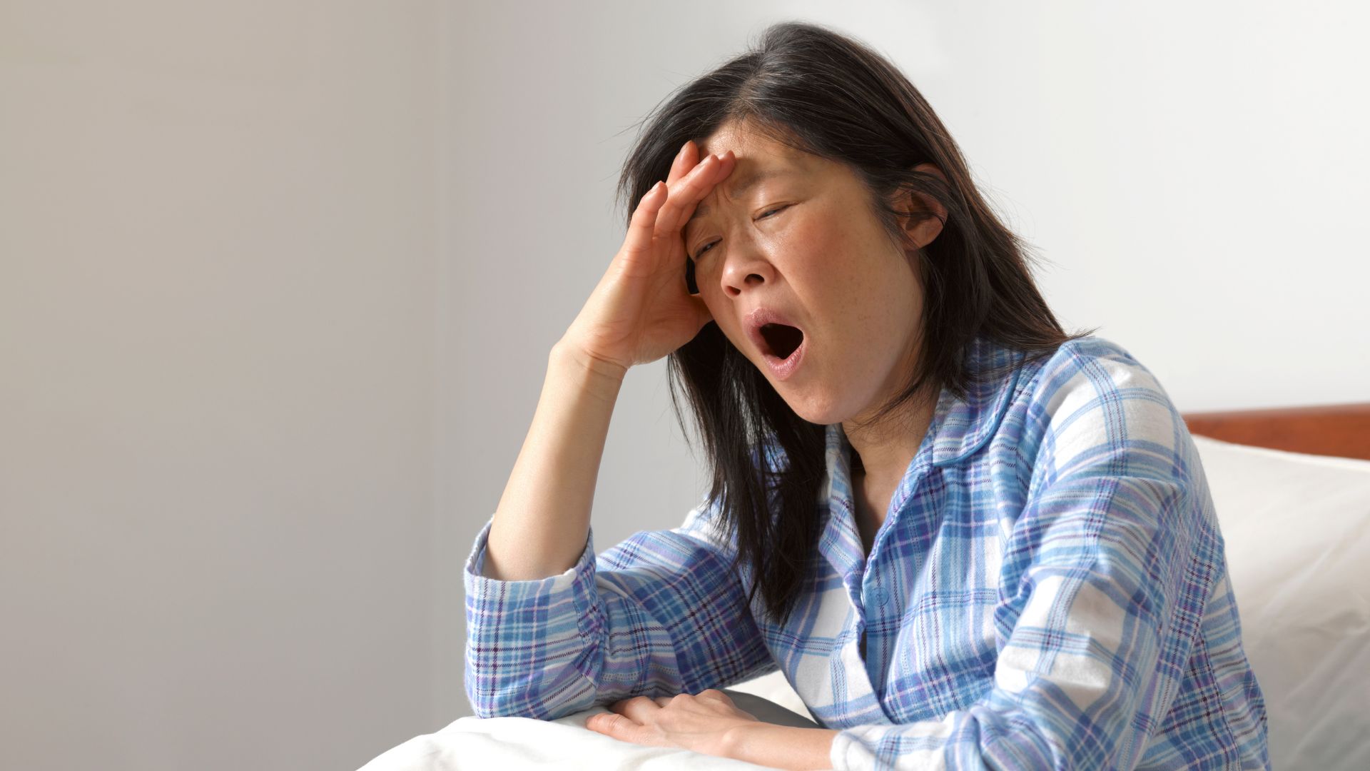 نیلی اور سفید چیک شدہ پاجامہ قمیض پہنے ایک عورت اپنے بستر پر بیٹھی جمائی لے رہی ہے۔