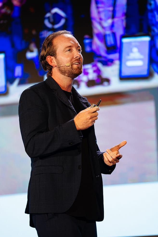 کالے سوٹ کی جیکٹ اور کالی قمیض پہنے ایک آدمی ریموٹ کنٹرول سے بات کر رہا ہے۔