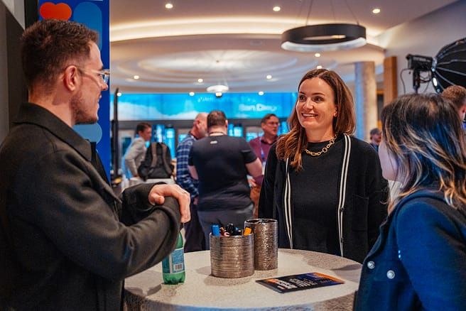 ایک مصروف کمرے میں، تین لوگ ایک اونچی میز کے گرد بیٹھے ہیں اور گفتگو کر رہے ہیں۔ بائیں طرف عینک پہنے ایک آدمی ہے، اور دائیں طرف دو عورتوں کو دیکھ رہا ہے۔ ایک عورت مسکرا رہی ہے اور دوسری اسے دیکھ رہی ہے۔