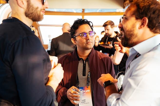 تین آدمی بات چیت کر رہے ہیں، دو درمیان میں موجود آدمی پر توجہ مرکوز کر رہے ہیں۔ درمیان میں موجود آدمی نے چشمہ پہنا ہوا ہے اور شراب پی رہا ہے۔