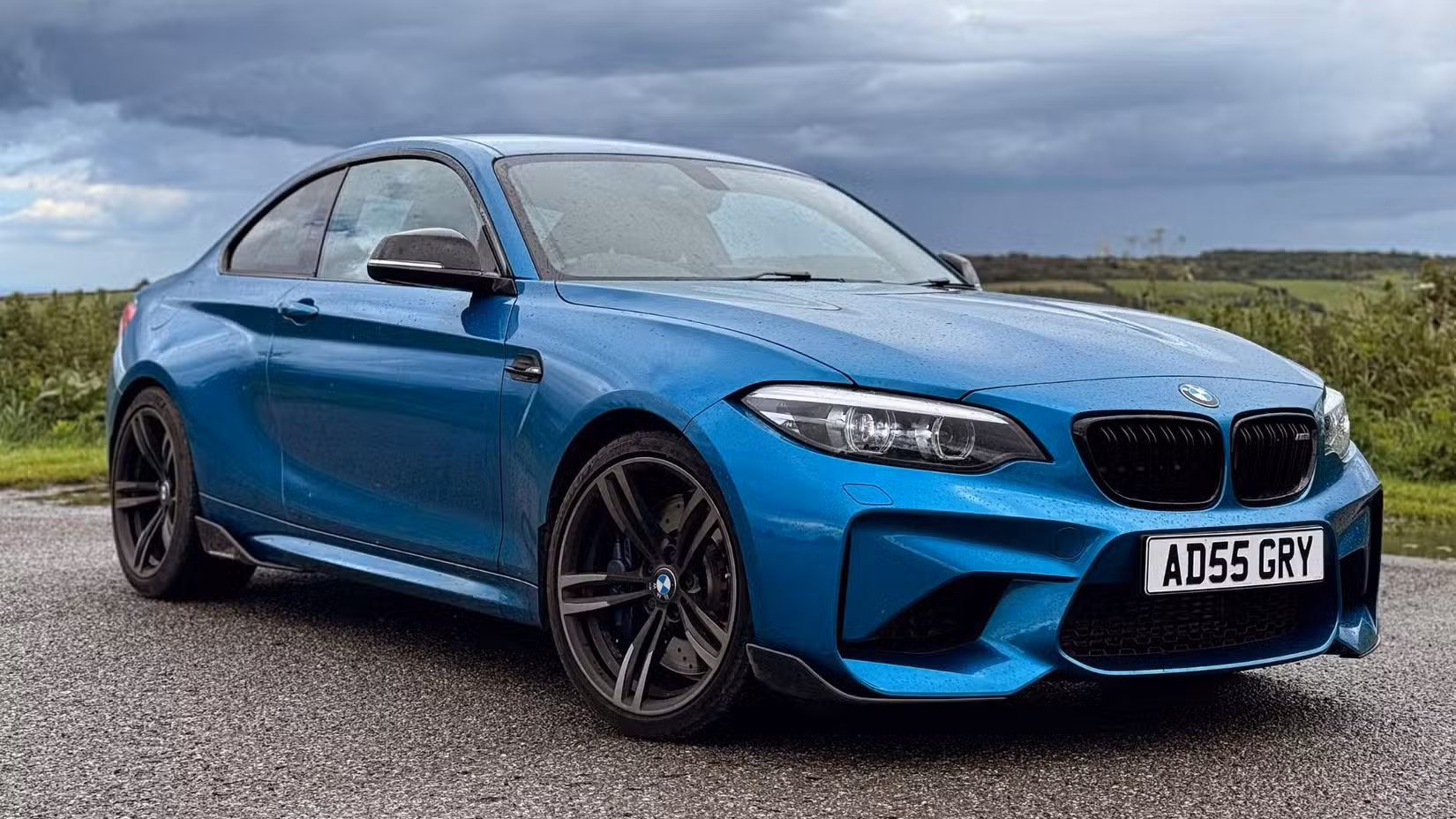 پس منظر میں فیلڈ کے ساتھ نیلے 2018 BMW M2 کا جامد سامنے والا 3/4 شاٹ۔
