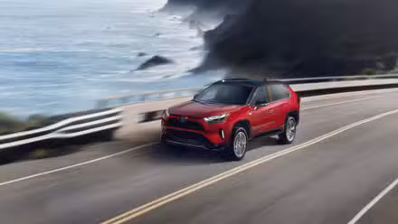 سرخ 2024 ٹویوٹا RAV4 Prime XSE کا متحرک سامنے والا 3/4 شاٹ سمندر کے ساتھ والی سڑک پر چلا رہا ہے۔