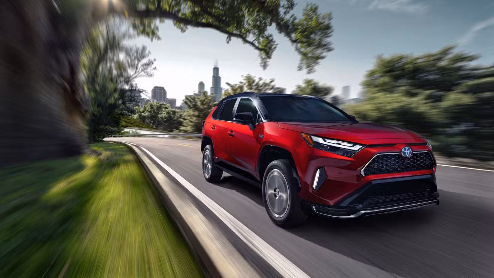 سرخ 2024 ٹویوٹا RAV4 پرائم XSE کا متحرک سامنے کا تین چوتھائی شاٹ پس منظر میں شہر کے منظر کے ساتھ درختوں کی قطار والی سڑک سے نیچے چلا رہا ہے۔